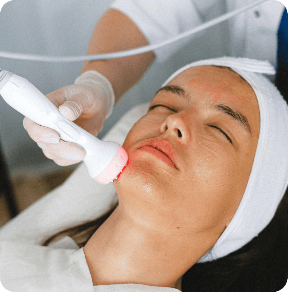 A person receiving a facial treatment with a device emitting red light while lying down with a headband, attended by someone wearing gloves, showcases an approach that bridges beauty care and primary care.