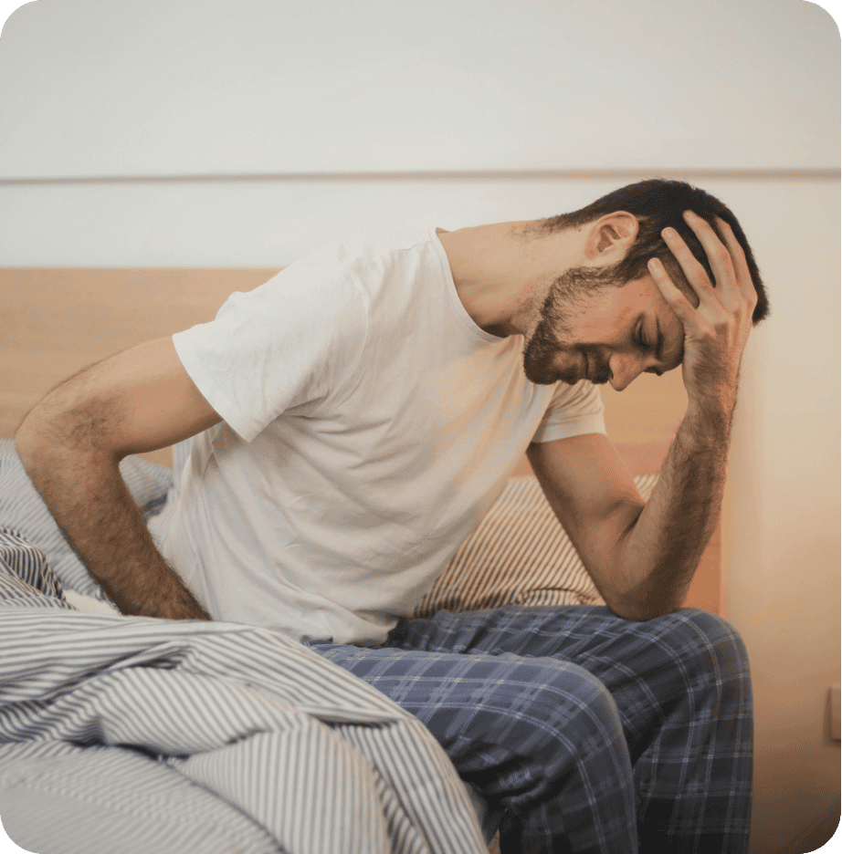 A man sits on a bed holding his head with one hand and his lower back with the other, appearing to be in pain. Wearing a white shirt and blue plaid pajama pants, he might be considering a visit to Primary Care for relief.