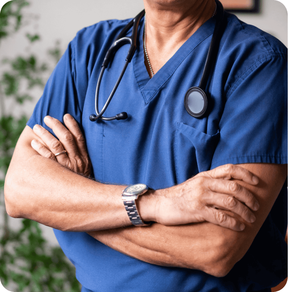 A person in blue medical scrubs with a stethoscope around their neck stands with arms crossed, embodying the dedication of primary care professionals.