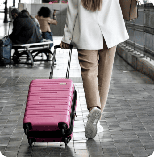 Person in a white coat, likely a primary care professional, pulling a pink suitcase with wheels, walking in an outdoor area with other people in the background.