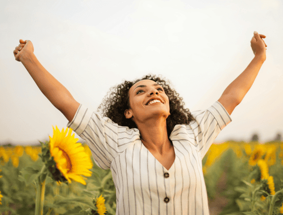 A person in a striped shirt stands in a sunflower field with arms raised and a joyful expression, like the feeling of relief after a visit to primary care.