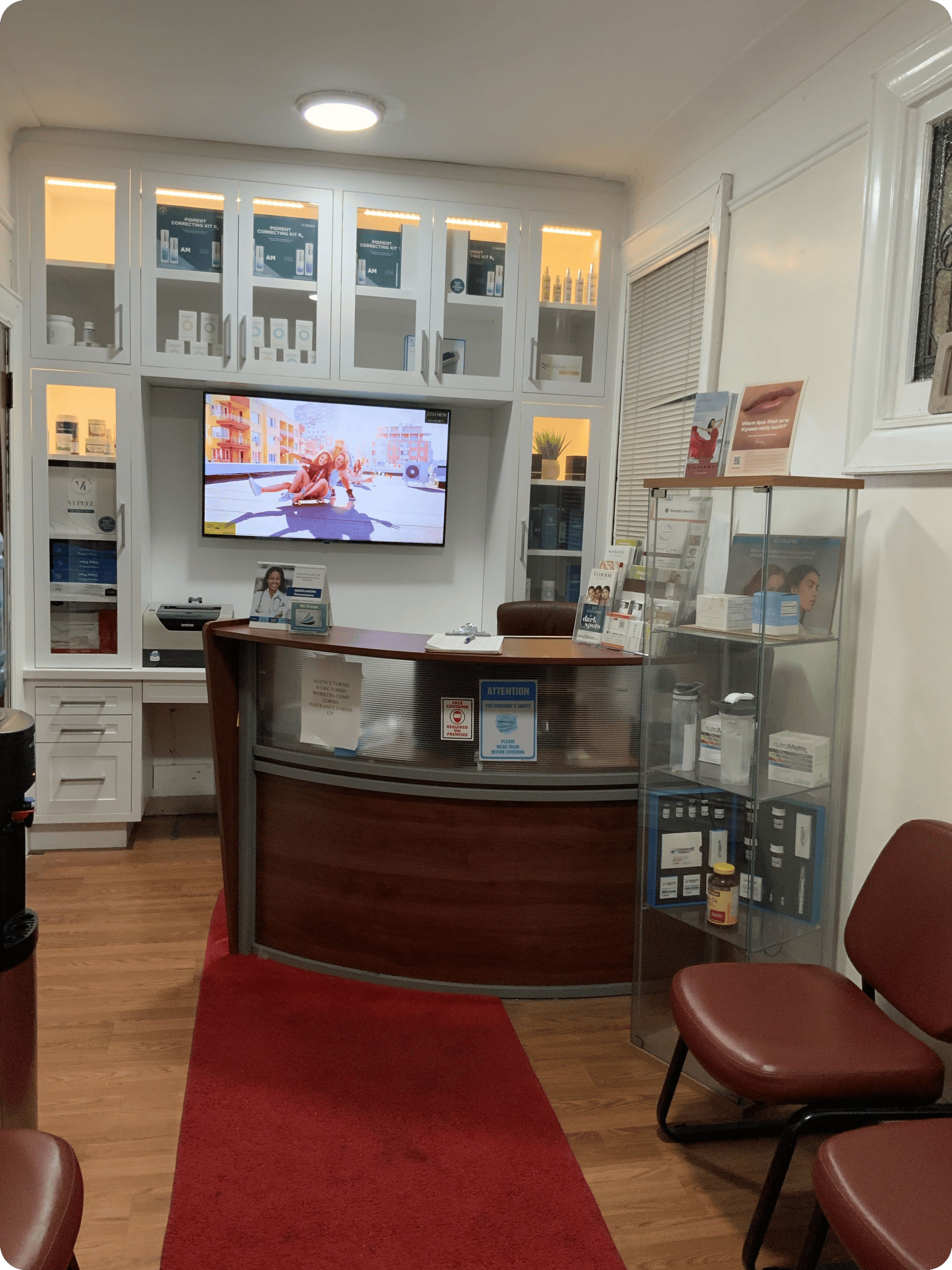 Small primary care office waiting area with a curved reception desk, glass display cabinets, wall-mounted TV showing a cartoon, and red cushioned chairs on a wooden floor.