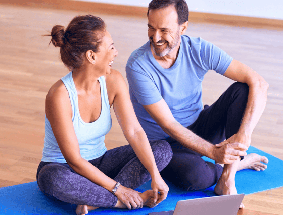 A man and a woman sit on a yoga mat, smiling at each other in a well-lit room with wooden flooring, embodying the essence of well-rounded primary care.
