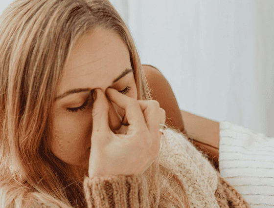 A woman with closed eyes is sitting and pinching the bridge of her nose, appearing to be in discomfort, possibly waiting for primary care. She has light hair and is wearing a sweater.