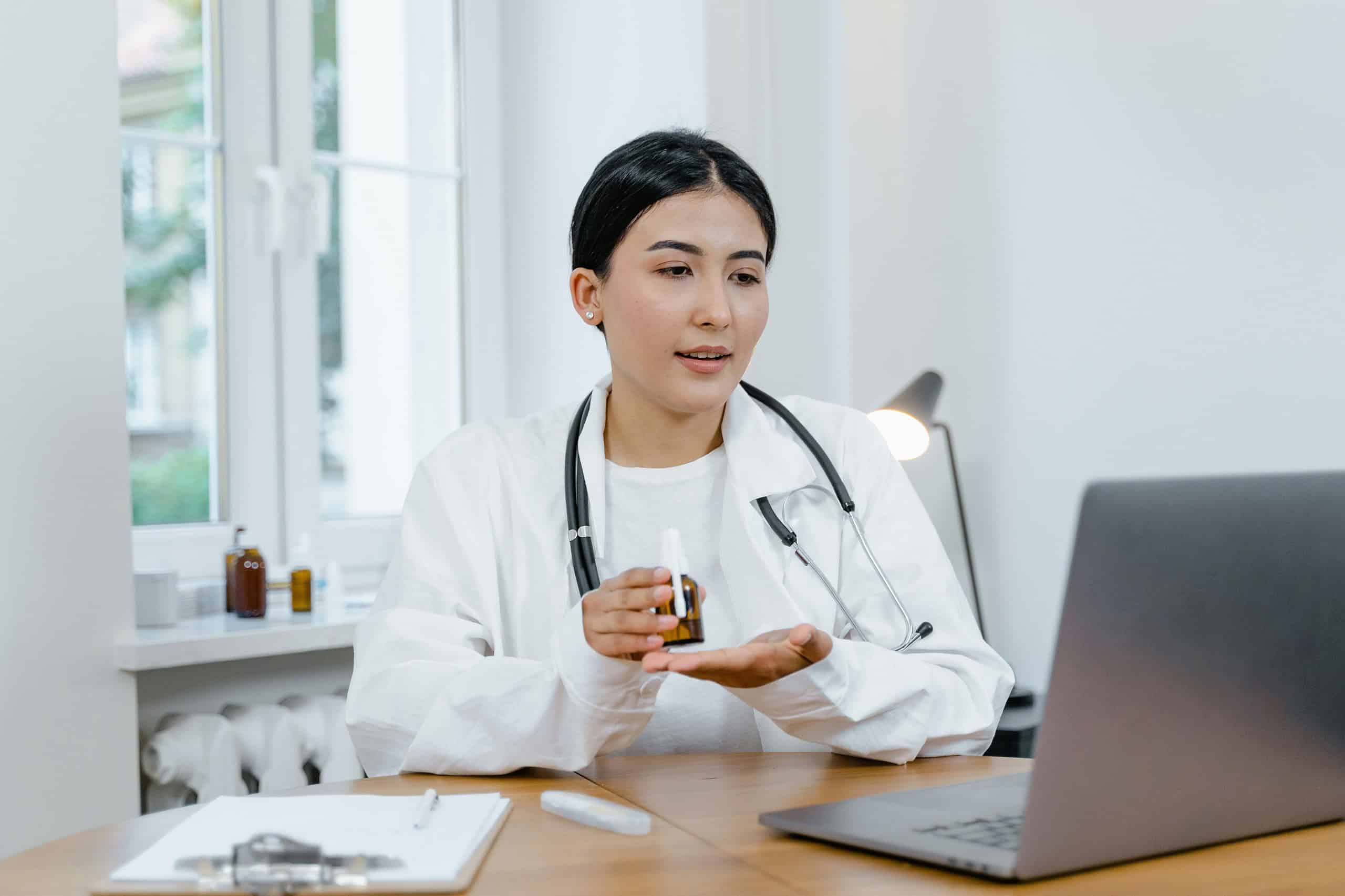 A primary care doctor in a white coat and stethoscope appears engaged in a video call, holding a small medication bottle in one hand, sitting at a desk with a clipboard, pen, and laptop.