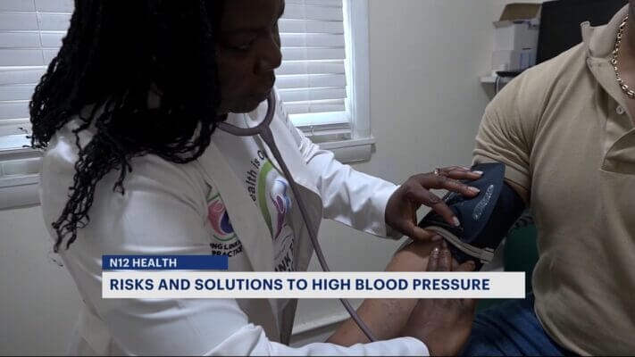 A healthcare professional measures a patient's blood pressure. Text on the image reads, "RISKS AND SOLUTIONS TO HIGH BLOOD PRESSURE.
